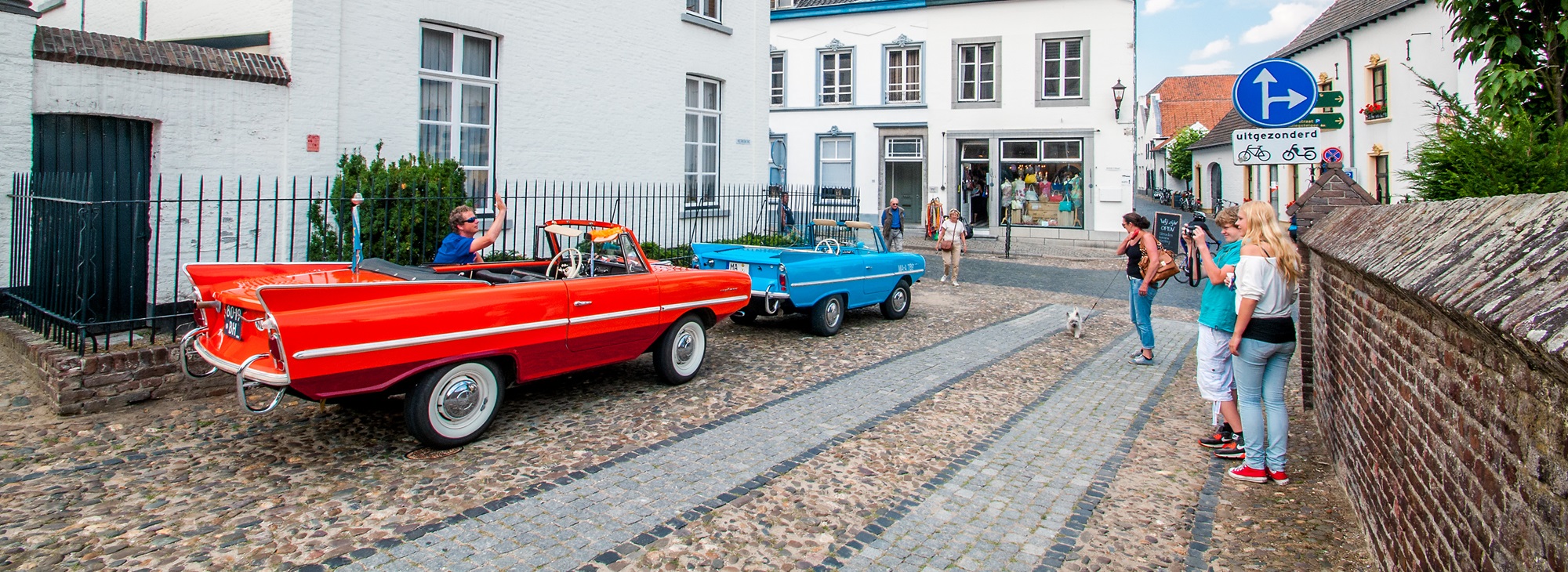 Twee kleurrijke oldtimers rijden over de kinderkopjes van een charmant historisch straatje, terwijl voorbijgangers foto's maken en genieten van de sfeer. Met de auto naar Limburg ontdek je zulke sfeervolle dorpen vol karakter en gezelligheid.