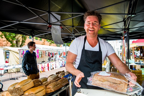 Lachende marktkramer op een weekmarkt in Limburg, die versgebakken brood aanbiedt onder een marktkraam.