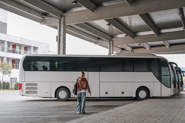 Woman with backpack stands in a covered car park looking at a parked coach. More buses and a modern building are visible in the background.