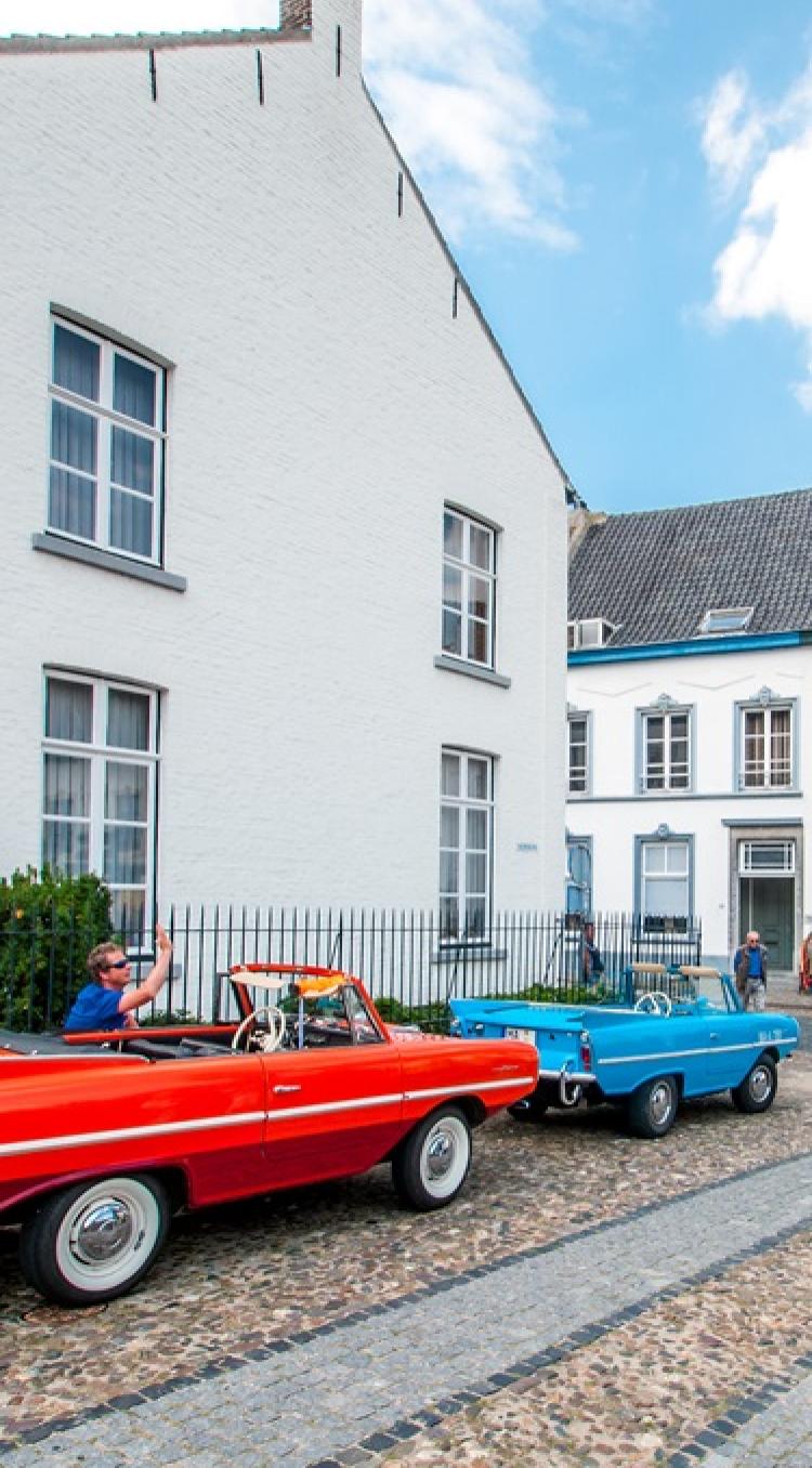 Twee kleurrijke oldtimers rijden over de kinderkopjes van een charmant historisch straatje, terwijl voorbijgangers foto's maken en genieten van de sfeer. Met de auto naar Limburg ontdek je zulke sfeervolle dorpen vol karakter en gezelligheid.
