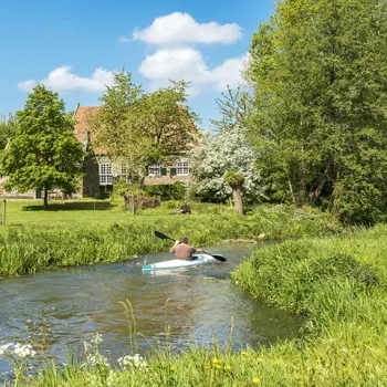 Persoon in een kano peddelt door een smalle rivier omringd door groen, met een historische boerderij op de achtergrond.