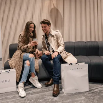 Man en vrouw zitten op een bank naar een telefoon te kijken. Er staan winkeltassen langs hun